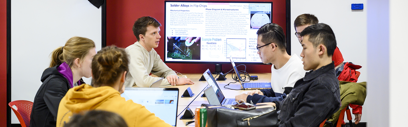 Students sitting around a table with their laptops out, and a shared screen at the far end displaying the class topic of the day.